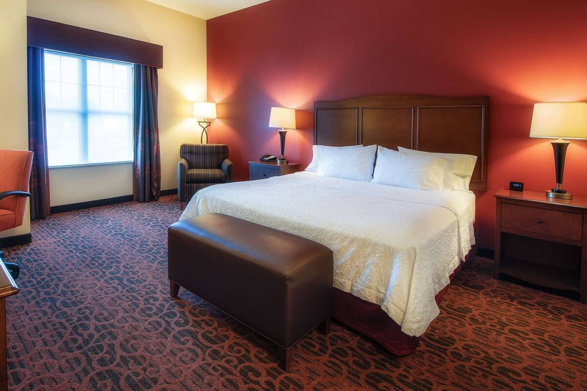 A hotel room with a large bed, two bedside lamps, a chair, a bench, and a nightstand with a phone.