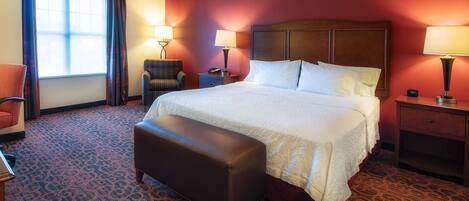 A hotel room with a large bed, two bedside lamps, a chair, a bench, and a nightstand with a phone.