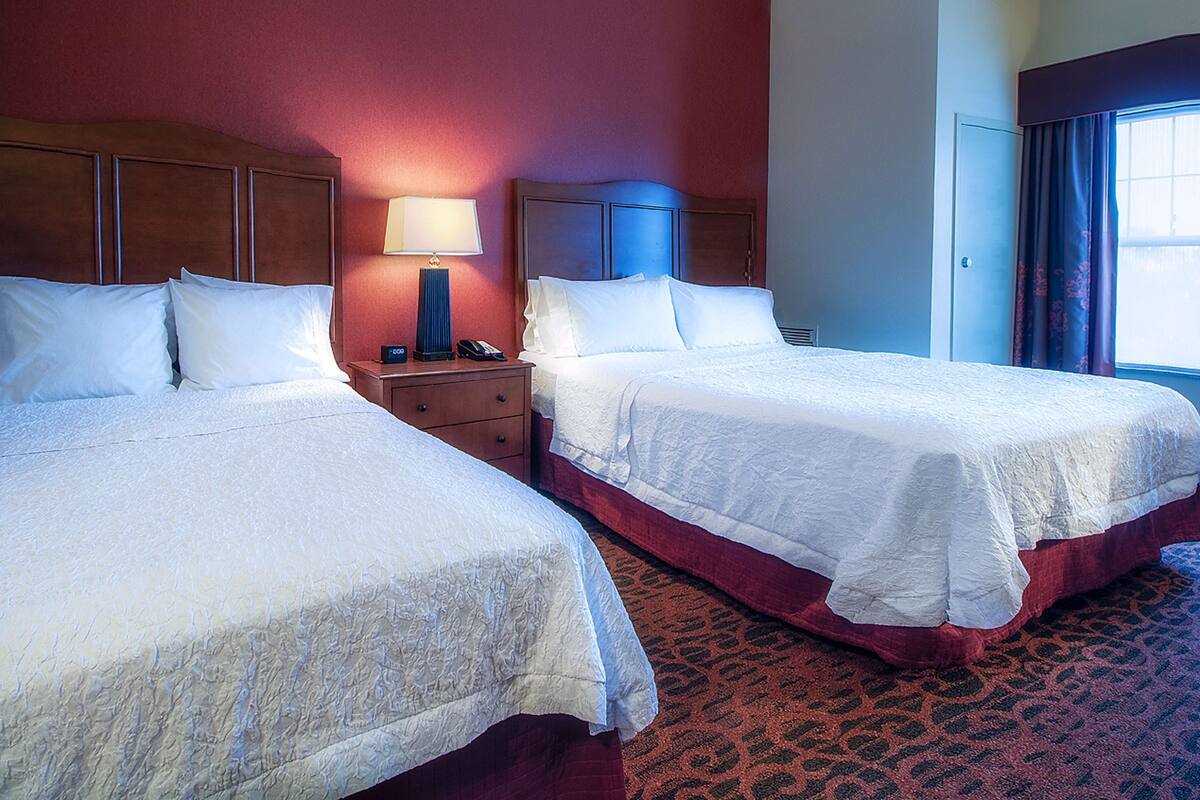 Two double beds in a hotel room with a wooden headboard, a nightstand with a lamp, and a patterned carpet.