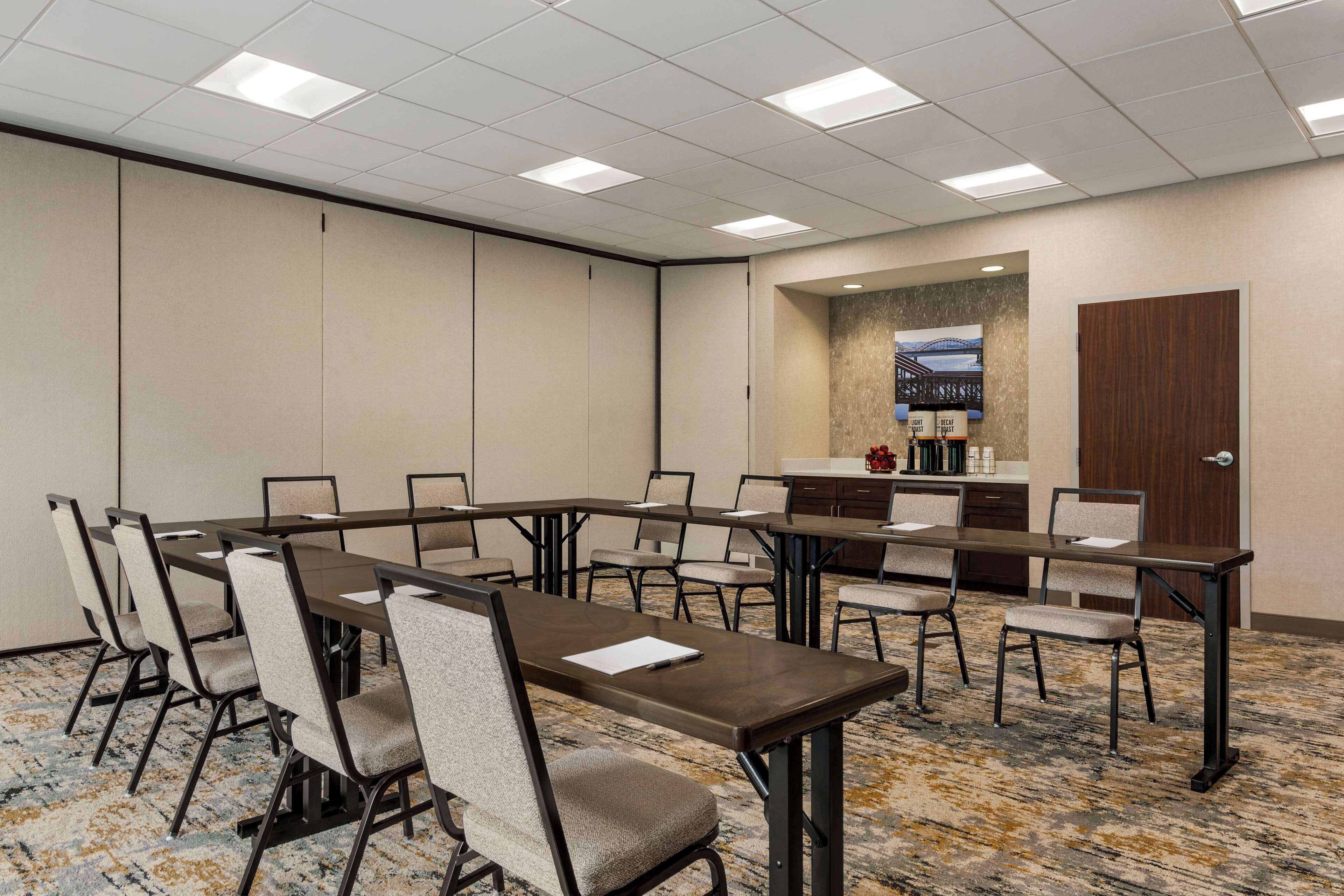 A conference room with long tables and chairs, a kitchenette, and a large window with a view.