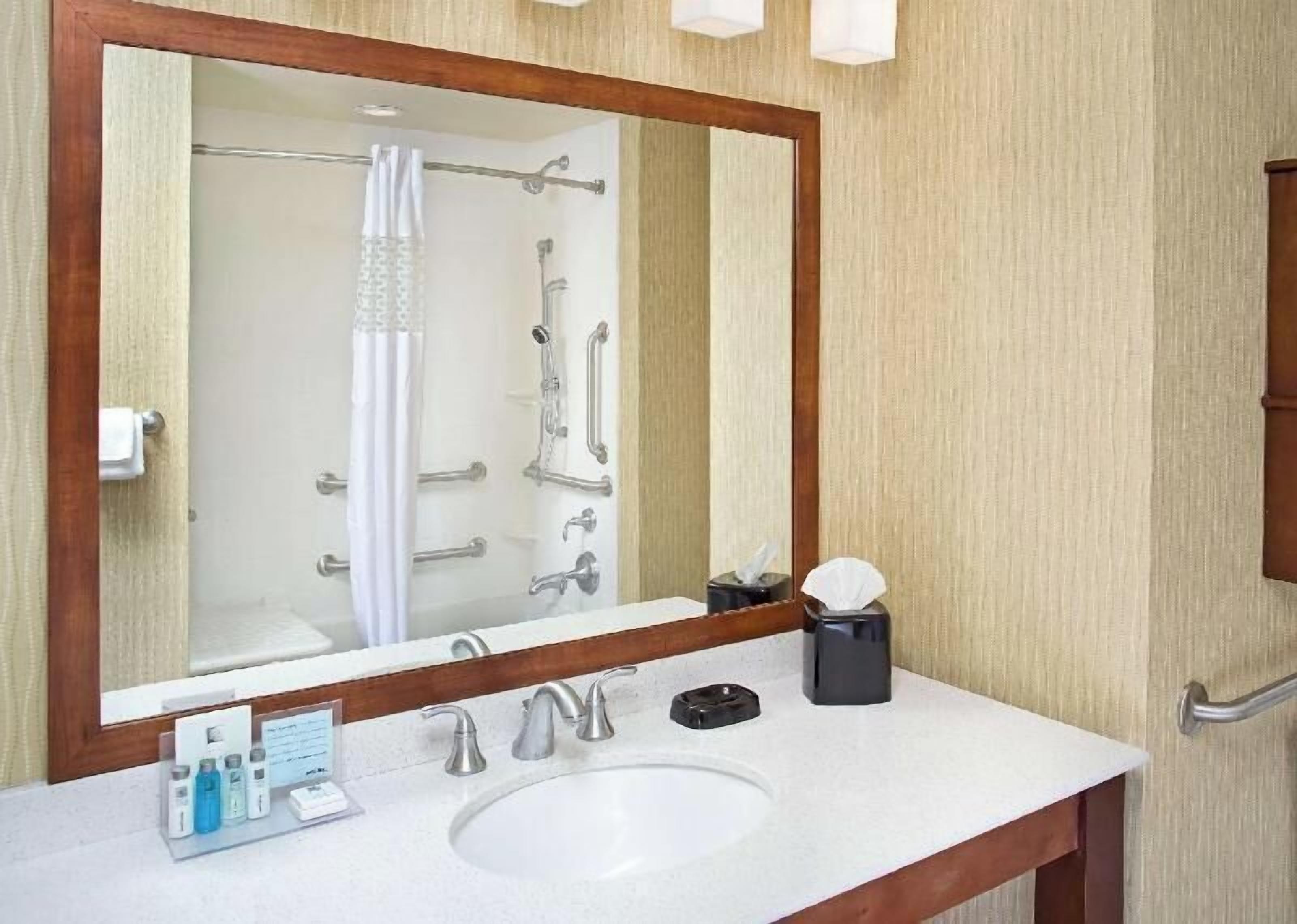 A bathroom with a large mirror, a sink, a shower with grab bars, and a towel rack.
