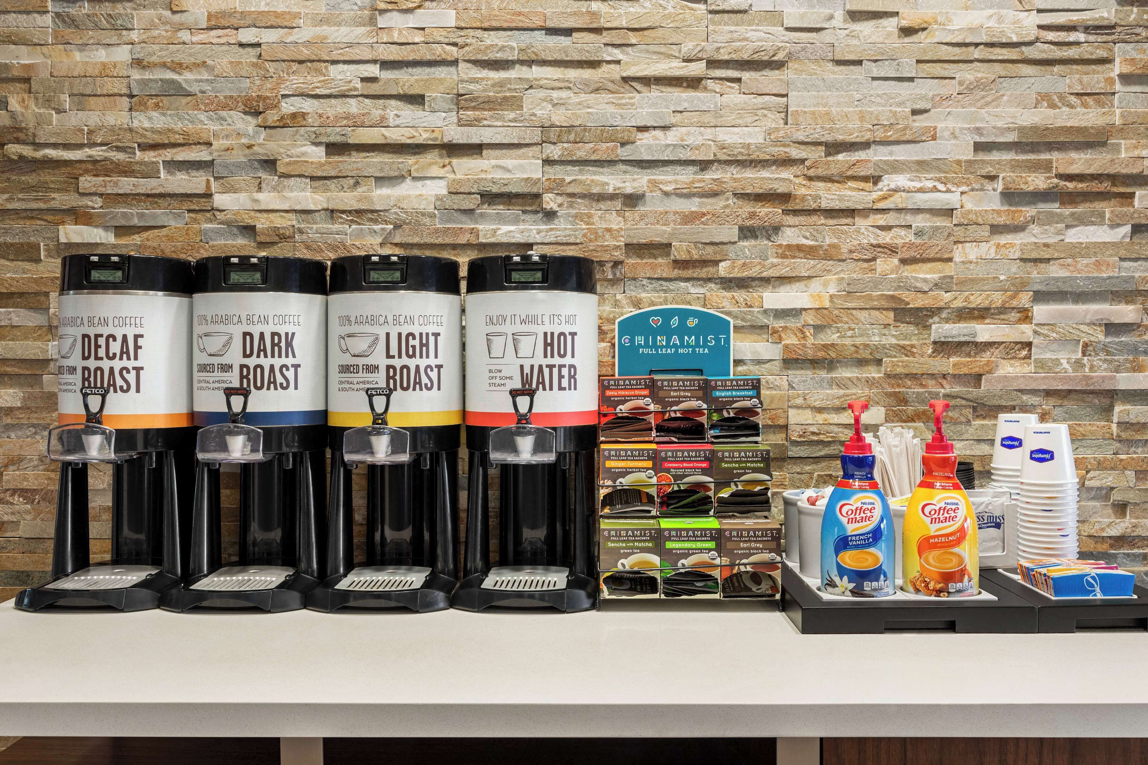 Cuatro dispensadores de café etiquetados como Descafeinado, Tueste Oscuro, Tueste Claro y Tueste Caliente, junto con un letrero para Chinamist té caliente de hojas sueltas.