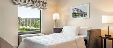 A hotel room with a large bed, a chair, a desk, a lamp, and a window with a view of buildings and trees.