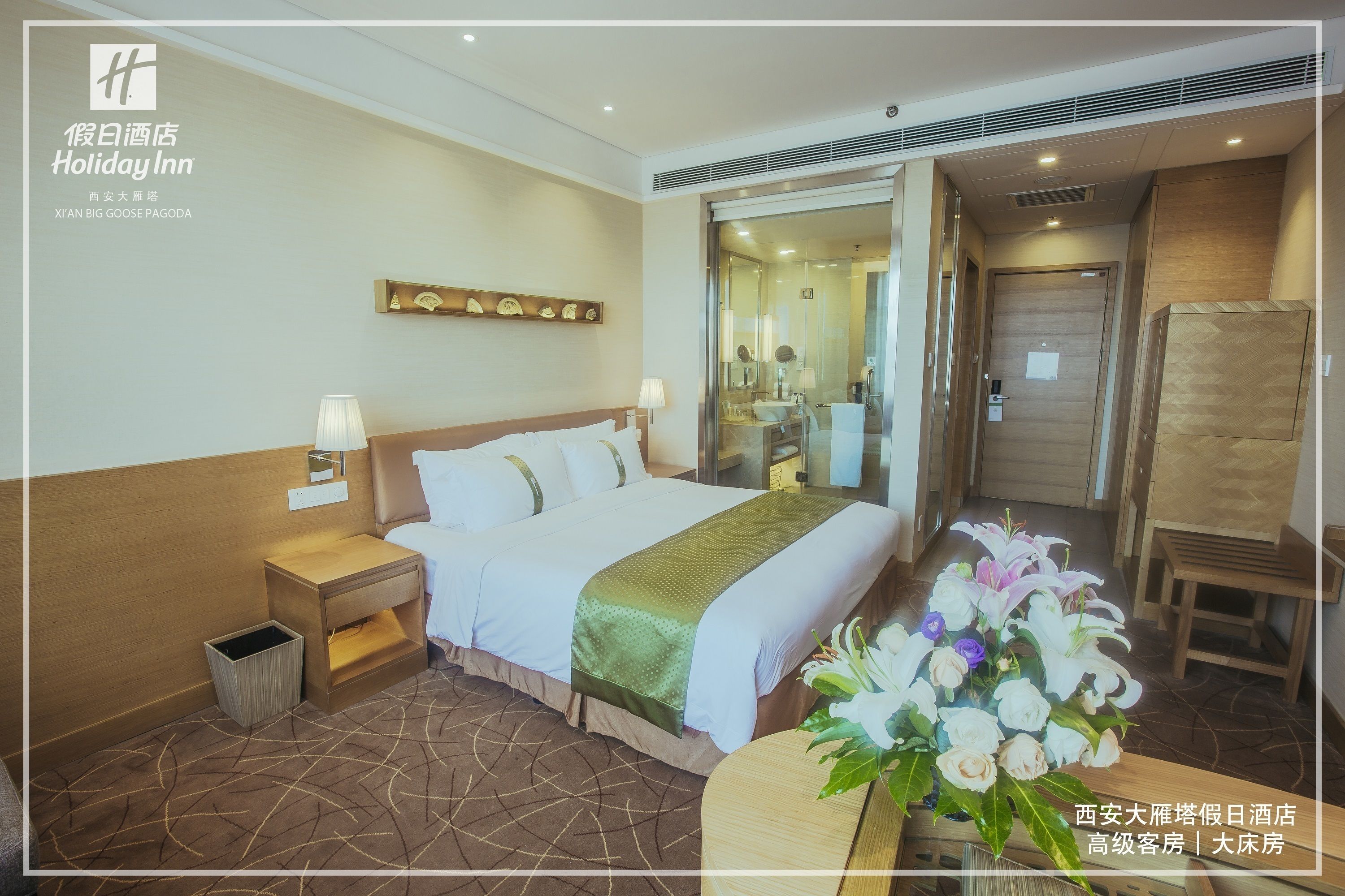 Une chambre d’hôtel avec un grand lit, des tables de chevet, un miroir, une tête de lit en bois, une petite table sur laquelle est posé un vase de fleurs, et une porte donnant sur la salle de bain.
