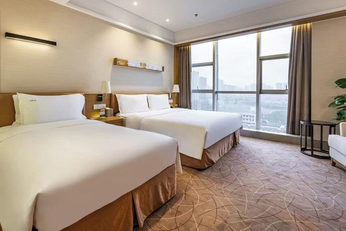 A hotel room with two beds, a sofa, a large window with a city view, and a potted plant.