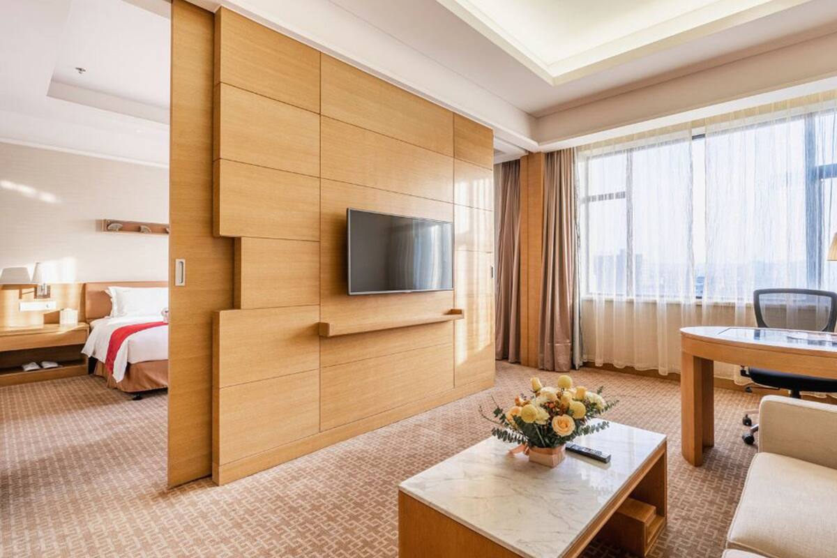 A hotel room with a bed, a desk, a chair, a TV mounted on the wall, and a flower vase on the coffee table.