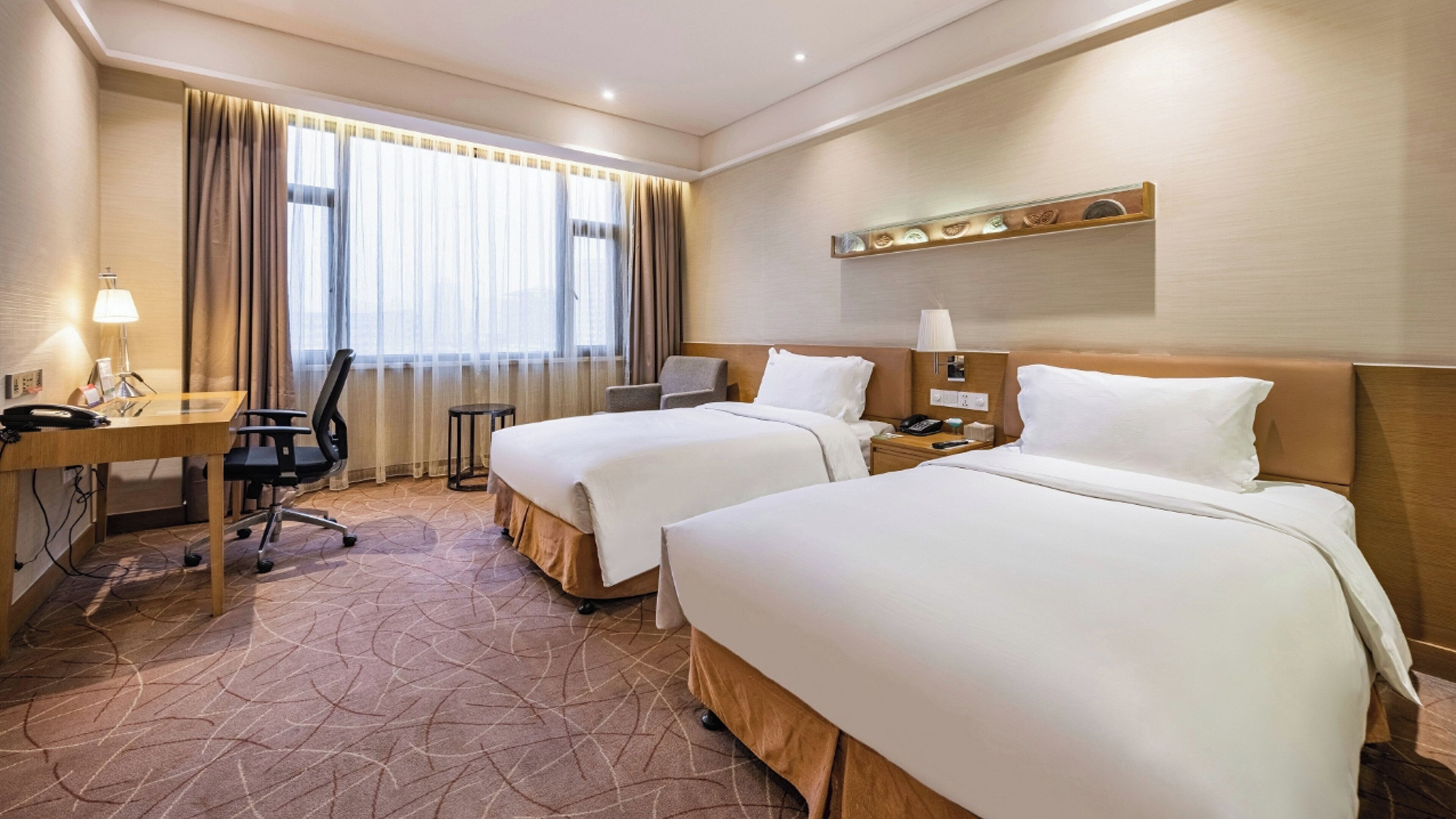 Ein Hotelzimmer mit zwei Betten, einem Schreibtisch mit Stuhl, einem kleinen Tisch und einem Fenster mit Vorhängen.