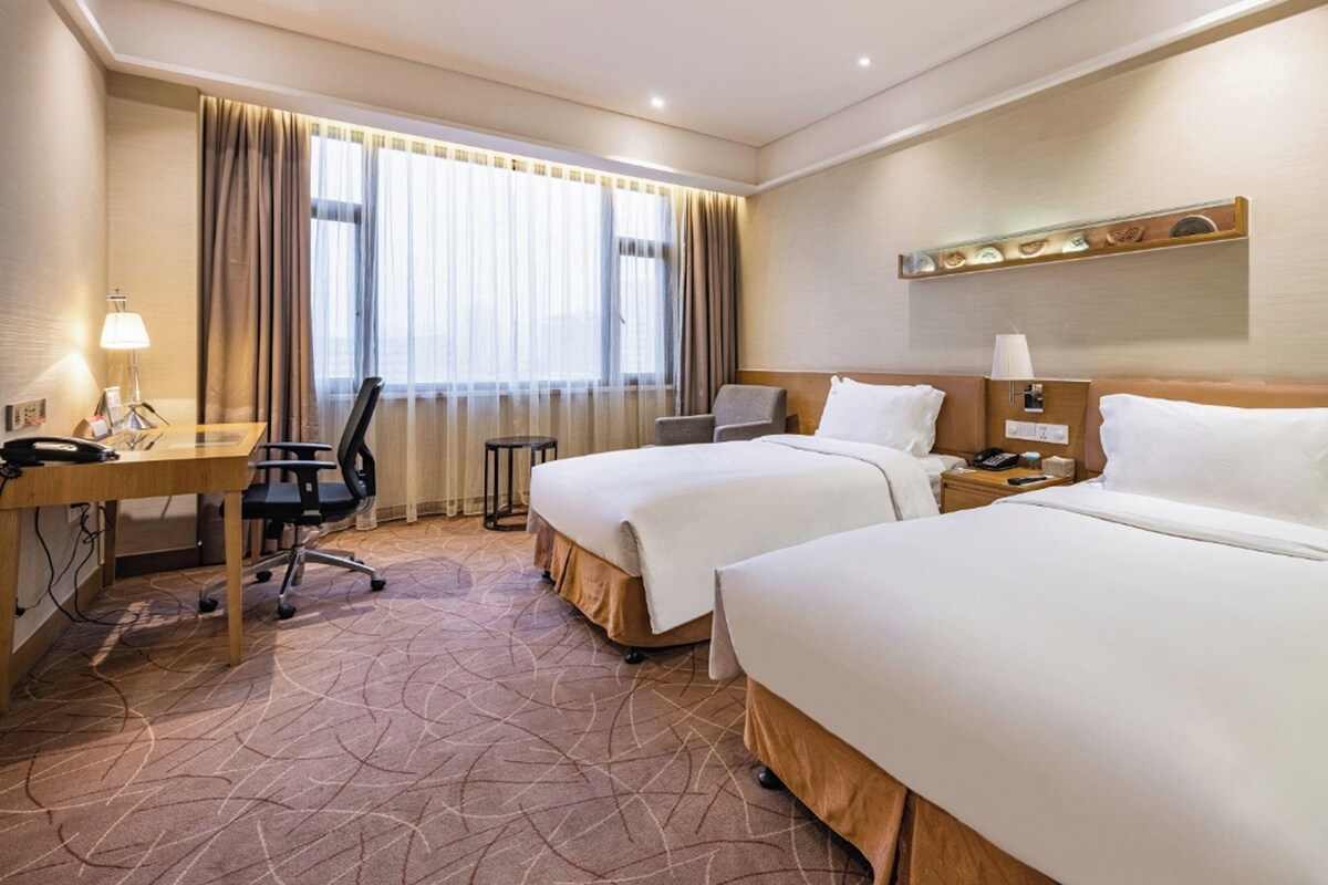 A hotel room with two beds, a desk with a chair, a small table, and a window with curtains.