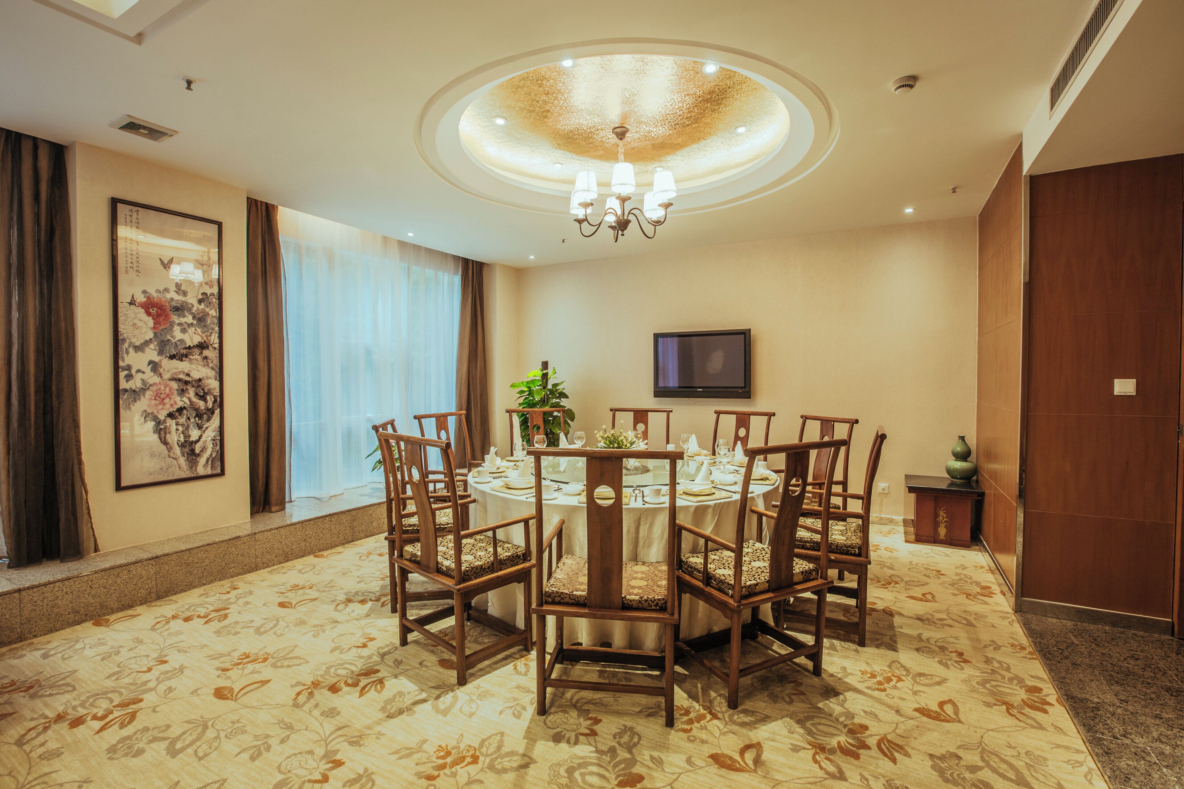 A dining area with a round table, wooden chairs, a chandelier, a painting, and a television on the wall.