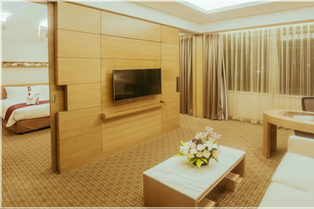 A hotel room with a flat-screen TV mounted on a wooden panel, a white sofa, a small coffee table with a floral arrangement, a desk with a lamp, and a bed with a red blanket.