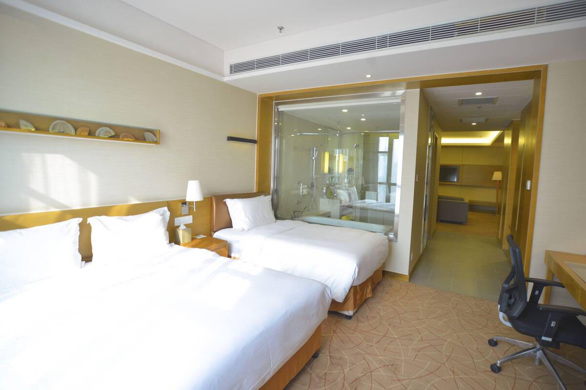 A hotel room with two beds, a bathroom with a glass shower, and a desk with a chair.