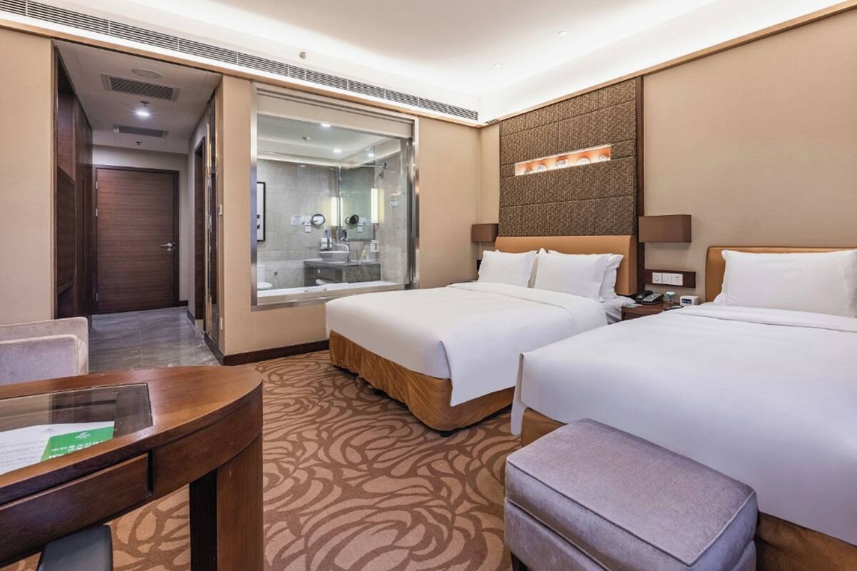 A hotel room with two beds, a desk, a chair, a bathroom with a glass shower enclosure, and a patterned carpet.