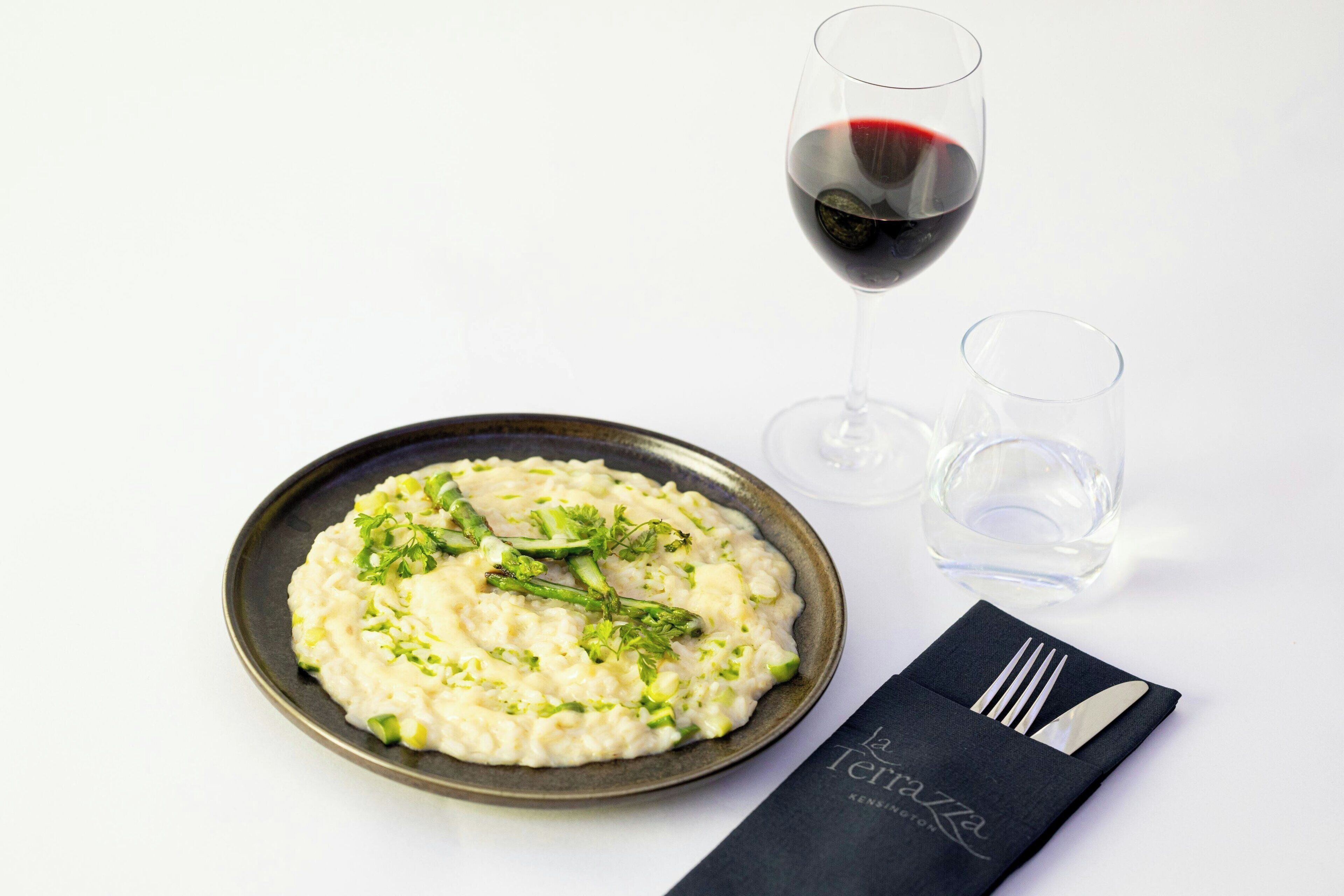 A plate of creamy risotto garnished with green herbs, a glass of red wine, and a folded napkin with a fork and knife on a white background.