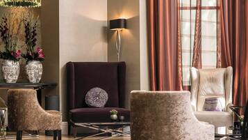 Ein modernes Wohnzimmer mit einem dunkelvioletten Sofa, zwei Sesseln und einer Stehlampe. Auf einem Beistelltisch stehen dekorative Vasen mit Blumen, und ein großes Fenster mit Vorhängen.