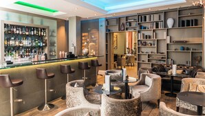 Un espace bar avec des tabourets, un comptoir et des étagères remplies de bouteilles. Il y a également des coins salon avec des chaises et des tables.