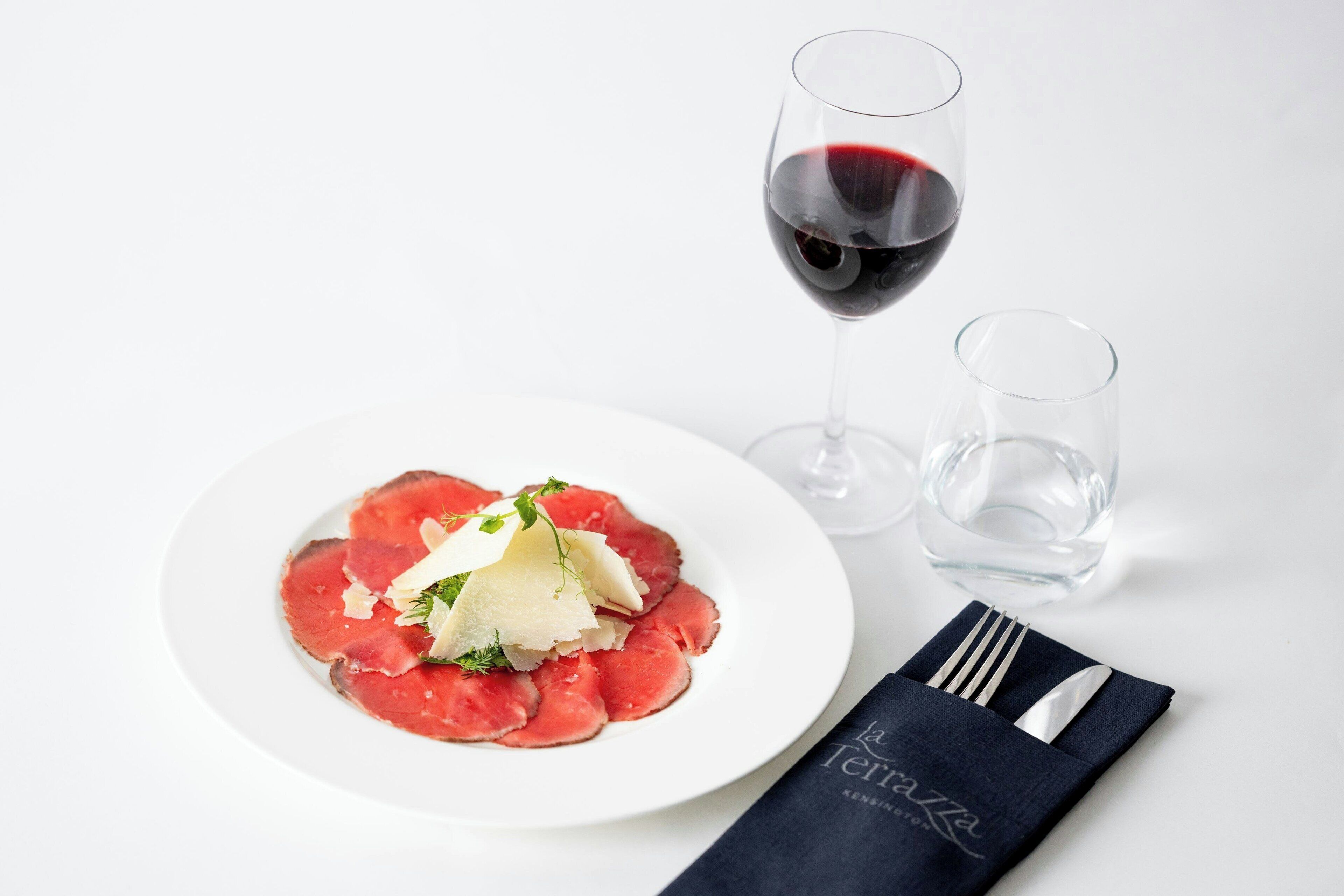 A white plate with a dish of sliced meat and cheese, a glass of red wine, and a napkin with a logo.
