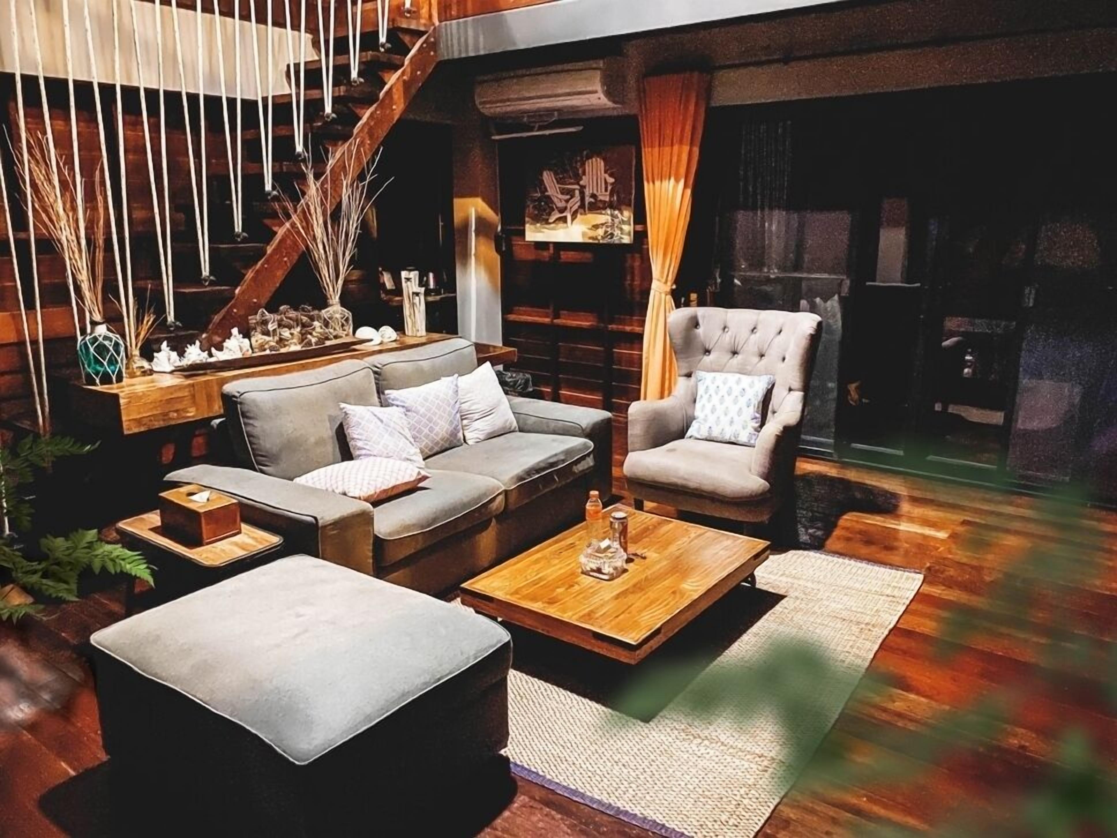 A cozy living room with a sofa, armchair, ottoman, and coffee table. A staircase with wooden railings and a fireplace are visible in the background.