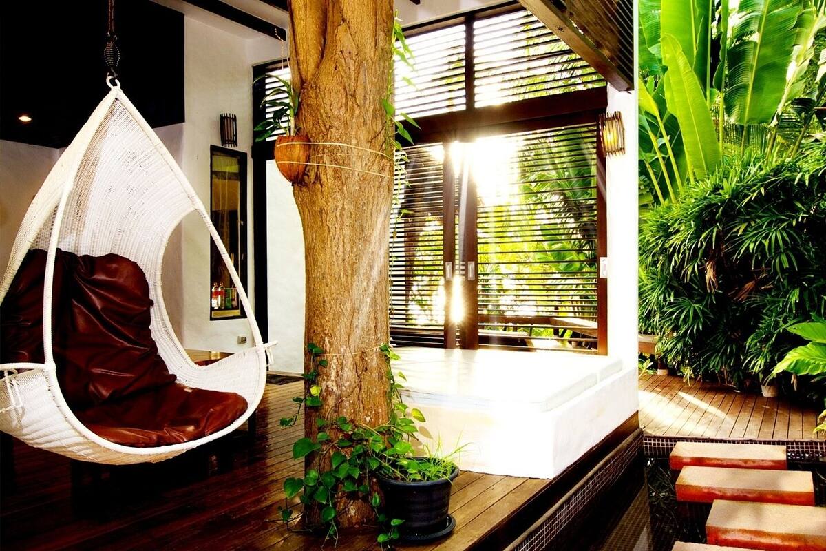 Un fauteuil suspendu dans une pièce moderne, avec un plancher en bois, un tronc d’arbre et une vue sur une végétation luxuriante à l’extérieur.