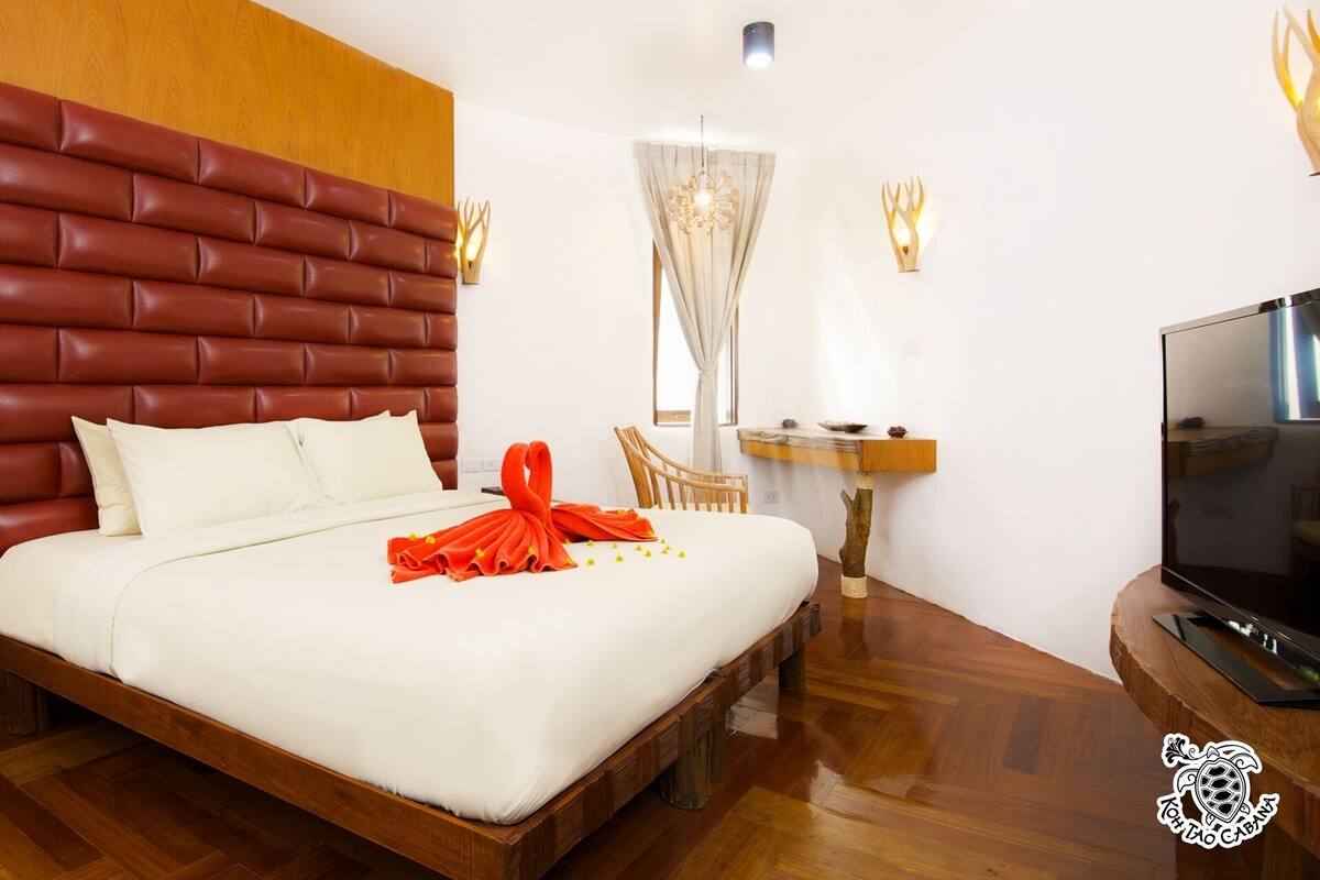 Une chambre d’hôtel avec une tête de lit en bois, un lit avec du linge de maison blanc, une serviette rouge, une chaise en bois, une petite table avec un bol, un téléviseur et un plancher en bois.