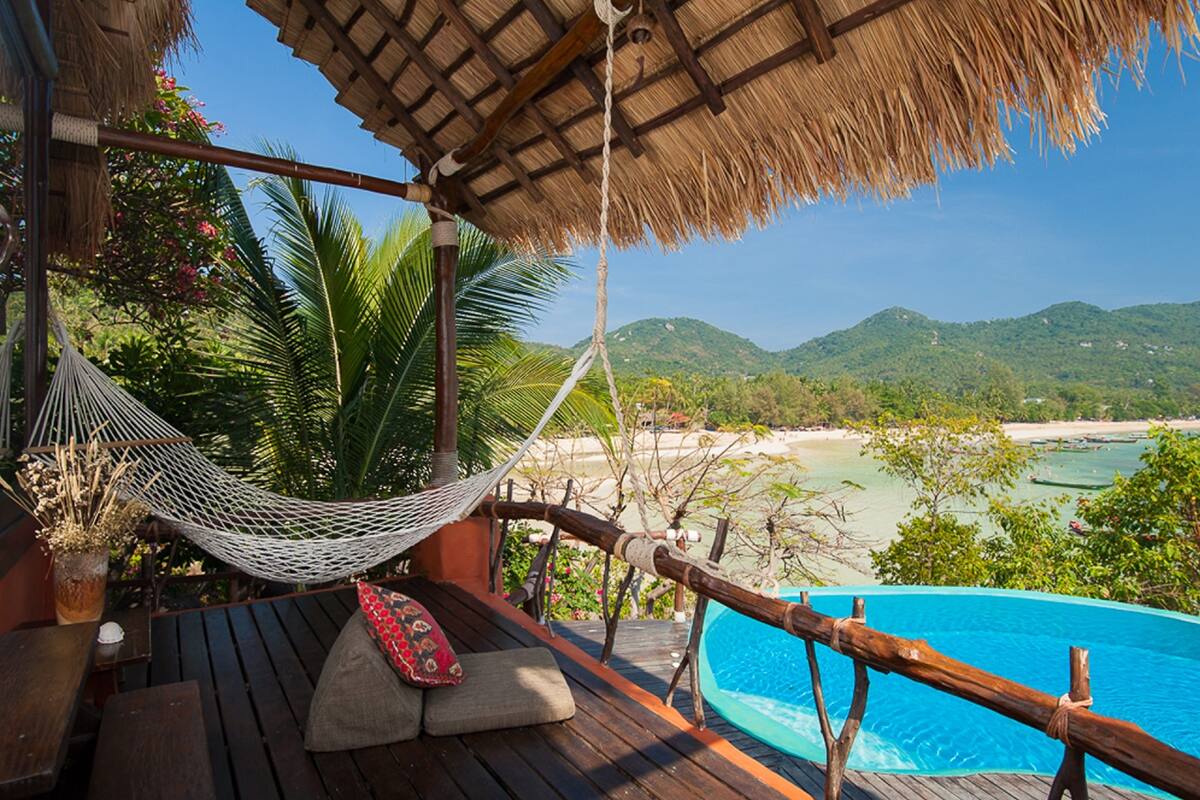 Une terrasse en bois avec un hamac, une piscine et une vue sur une plage et des montagnes.