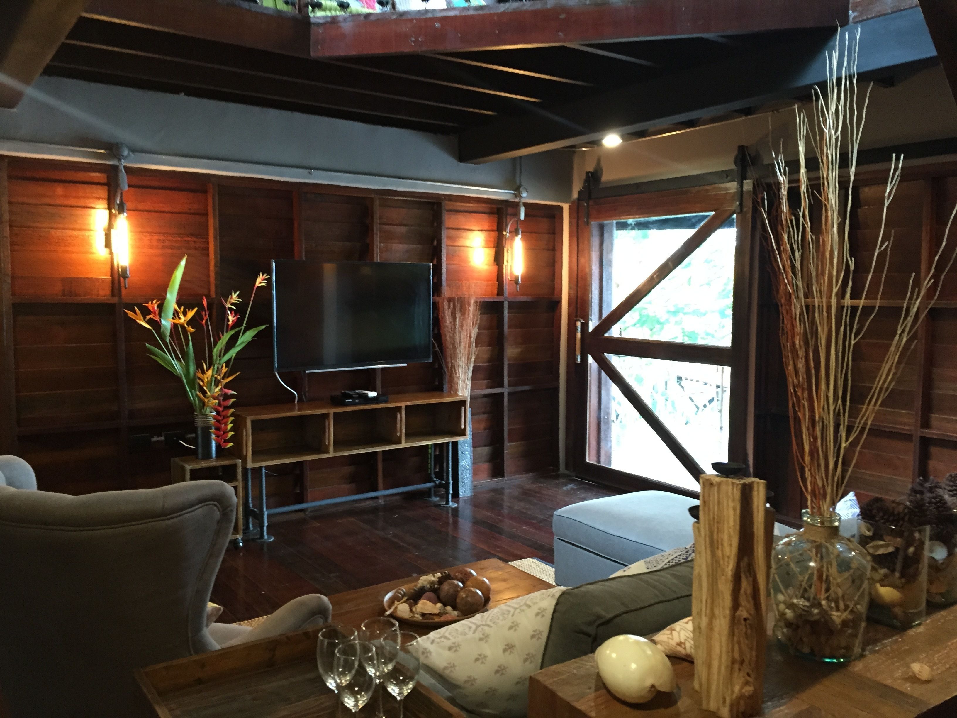 A cozy living room with a wooden TV stand, a flat-screen TV, a dining table with chairs, and a large window with a view of greenery.