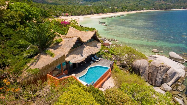 A thatched-roof structure with a pool, surrounded by lush greenery and overlooking a beach with boats.