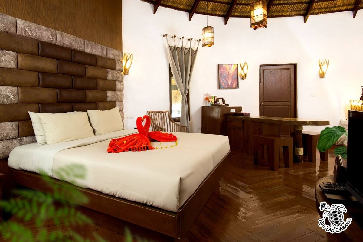 Une chambre d’hôtel avec une tête de lit en bois, un lit recouvert d’une courtepointe blanche, un bureau avec une chaise, une table à manger et un téléviseur.