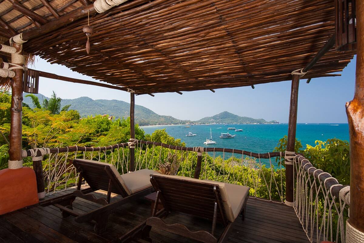 Une terrasse en bois avec deux chaises longues et un toit de chaume, offrant une vue sur une zone côtière avec des bateaux et de la végétation.