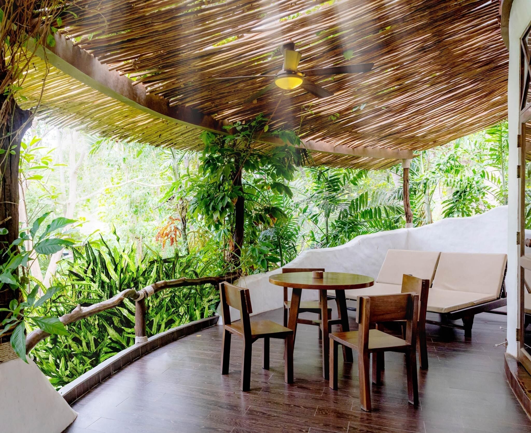 A covered outdoor seating area with a table and chairs, surrounded by lush greenery and a bamboo ceiling.