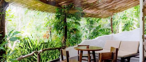 A covered outdoor seating area with a table and chairs, surrounded by lush greenery and a bamboo ceiling.