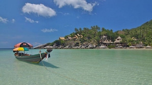 A clear turquoise sea with a boat anchored near the shore, a thatched-roof structure, and lush greenery on a hillside.