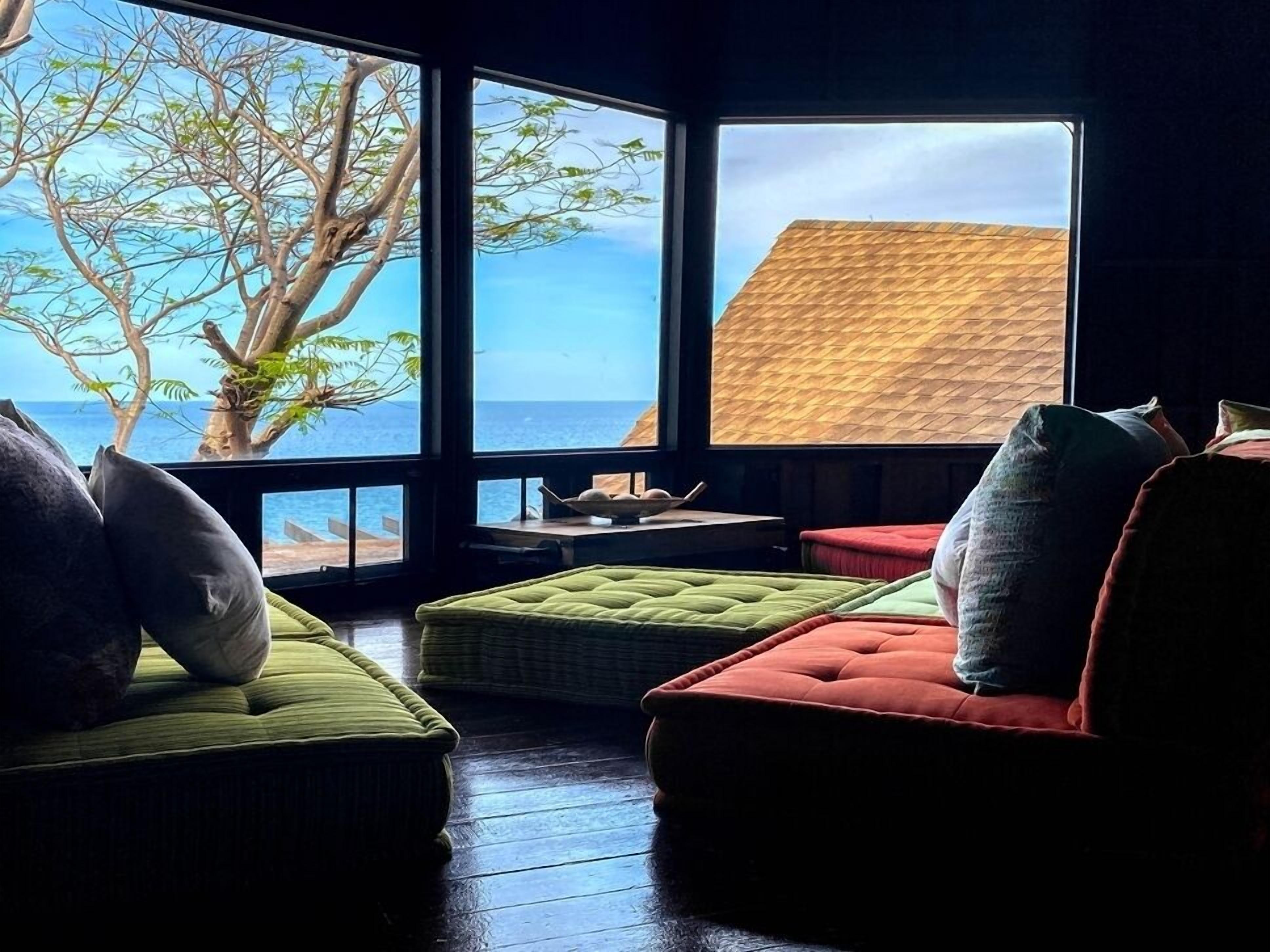 A room with large windows overlooking a beach and the sea. There are two green cushioned seating areas and a red cushioned seating area.