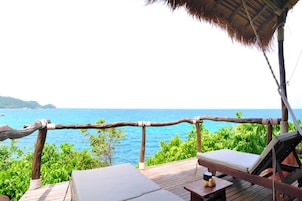 Terraza de madera con dos sillones de descanso y una mesa, con vista a un mar azul cristalino y exuberante vegetación.