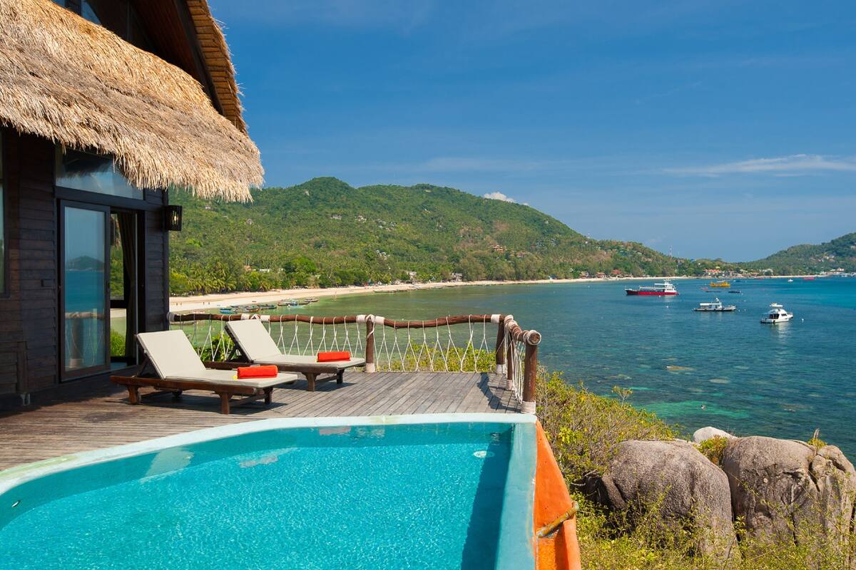 Un espace aménagé au bord de la piscine, avec des chaises longues, une terrasse en bois et une vue sur la mer et les collines.