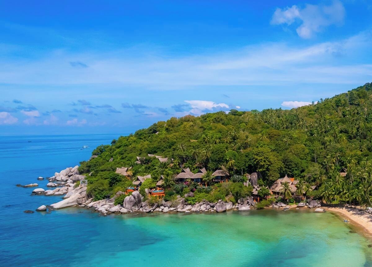 Un resort playero tropical con cabañas de techo de paja, una costa rocosa y aguas turquesas cristalinas.