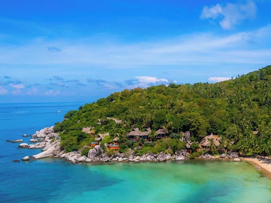 A tropical beach resort with thatched-roof cottages, a rocky shoreline, and clear turquoise waters.