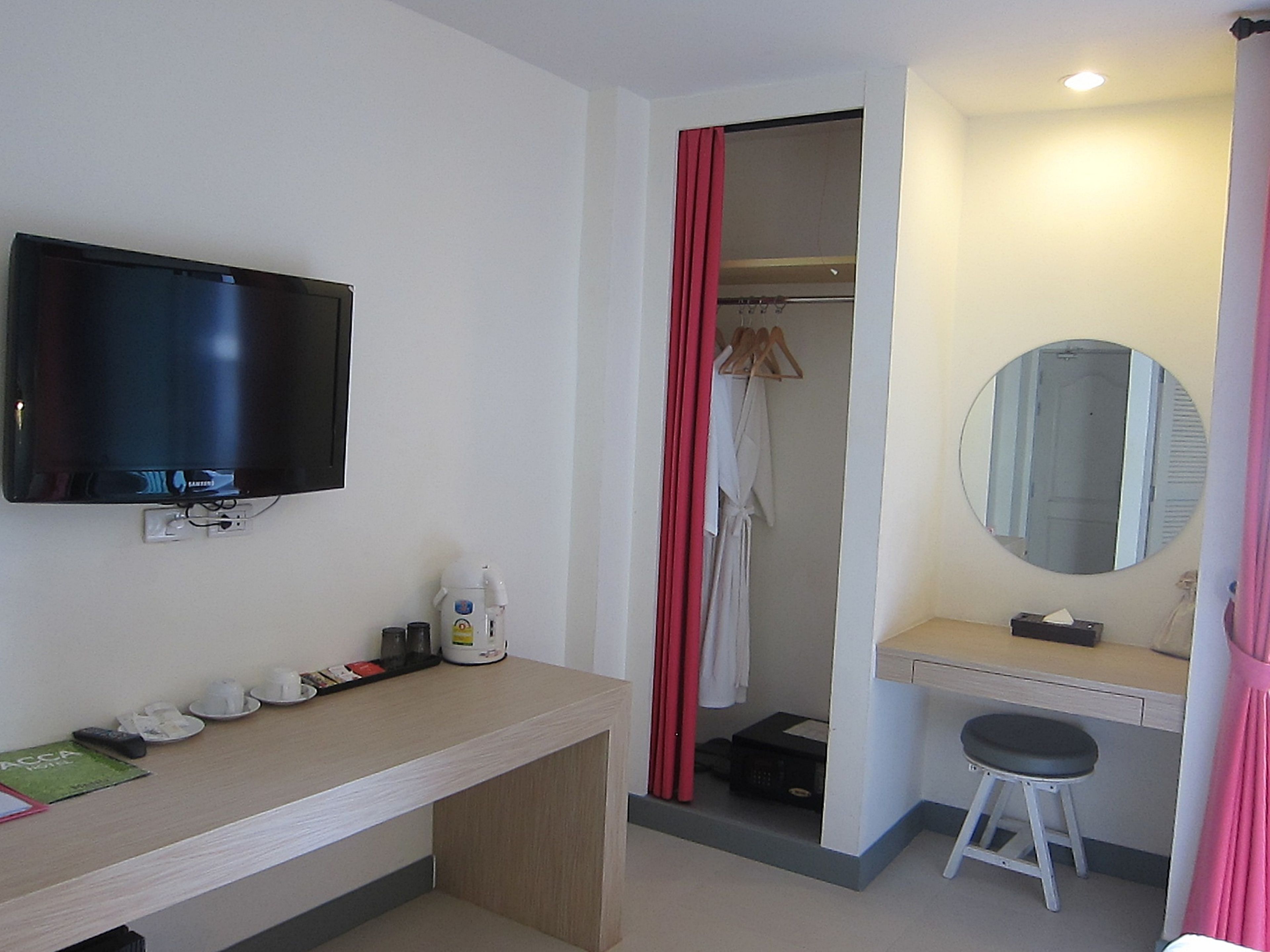 A hotel room with a flat-screen TV mounted on the wall, a wooden desk with a mirror, a small stool, and a shower area with a red curtain.