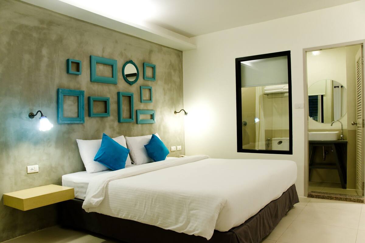 A modern bedroom with a large bed, bedside tables, a wall-mounted mirror, and a bathroom visible through an open door.