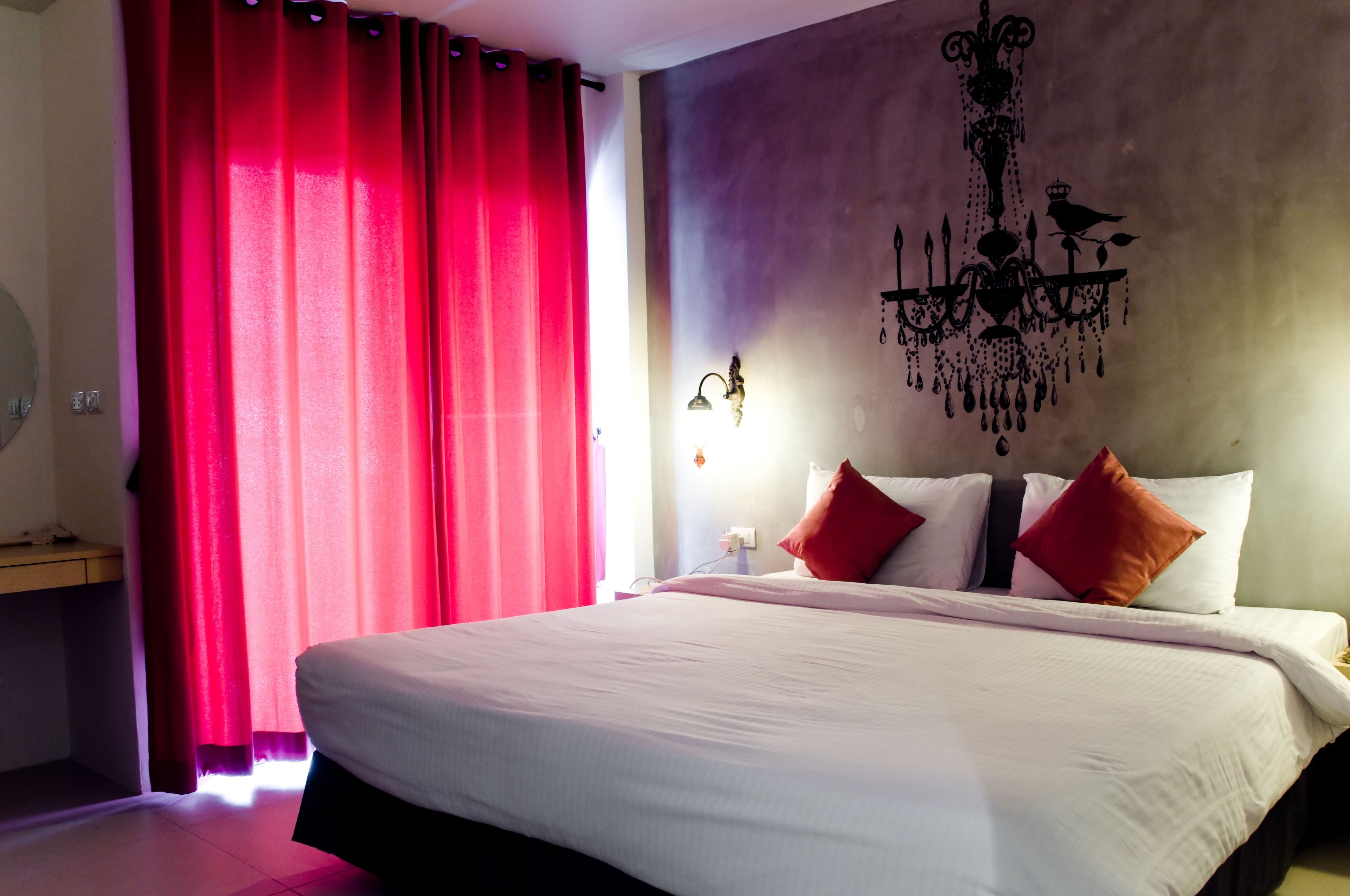 A hotel room with a large bed, red curtains, a chandelier, and a nightstand with a lamp.