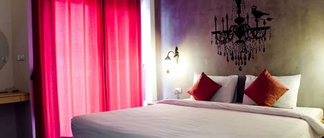 A hotel room with a large bed, red curtains, a chandelier, and a nightstand with a lamp.