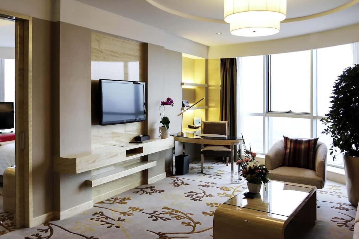 Ett modernt hotellrum med en platt-TV, ett skrivbord med stol, en soffa, ett soffbord och ett stort fönster.