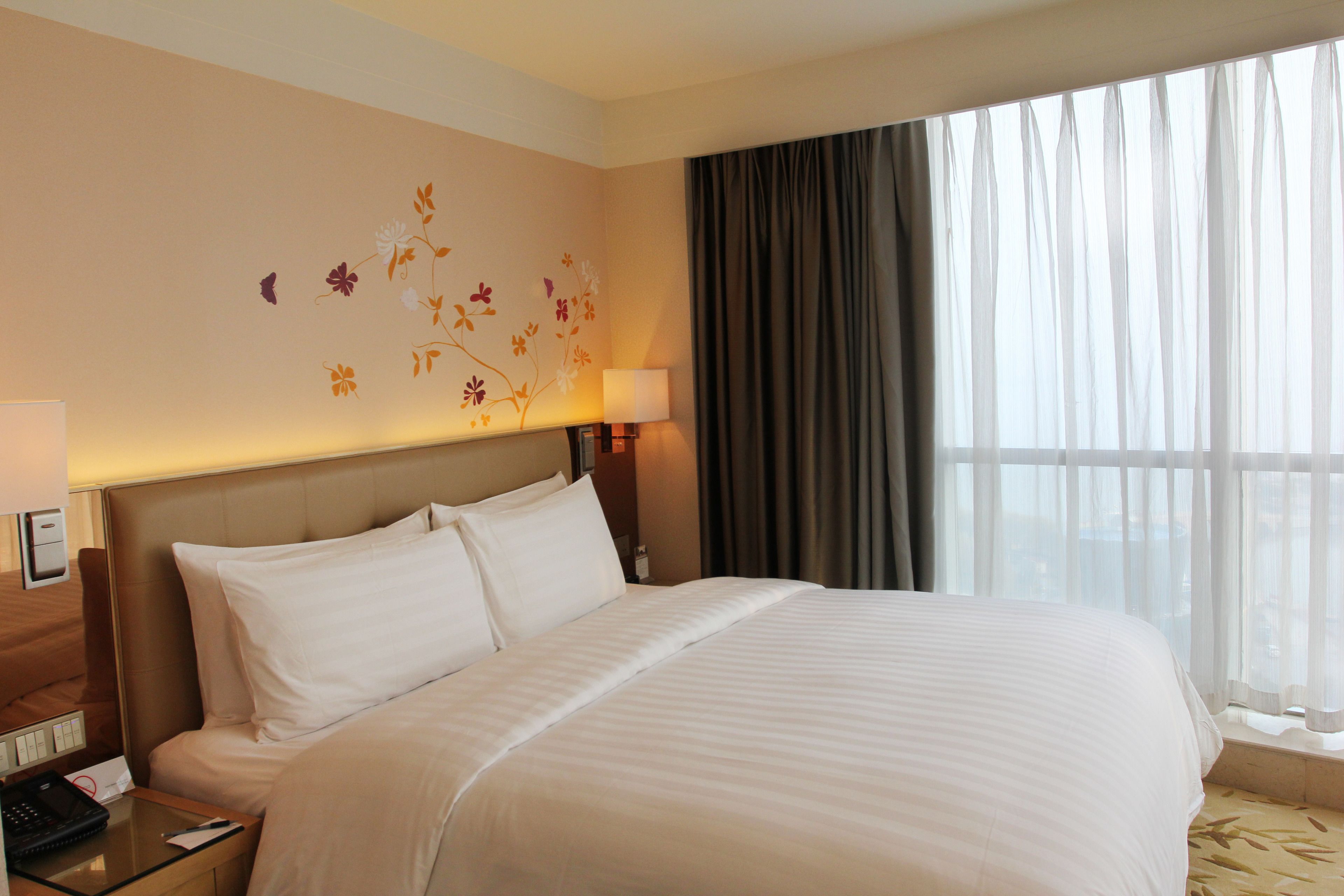 A hotel room with a large bed, a nightstand with a lamp, a floral wall decoration, and a window with curtains.