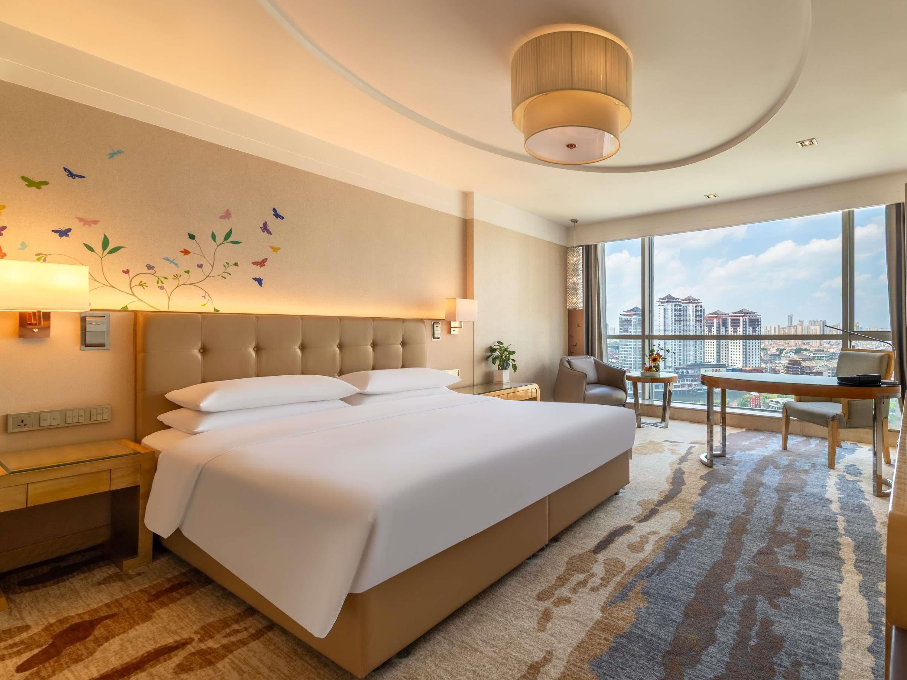 Quarto de hotel com cama grande, escrivaninha, cadeira e vista para a cidade, além de decoração na parede.