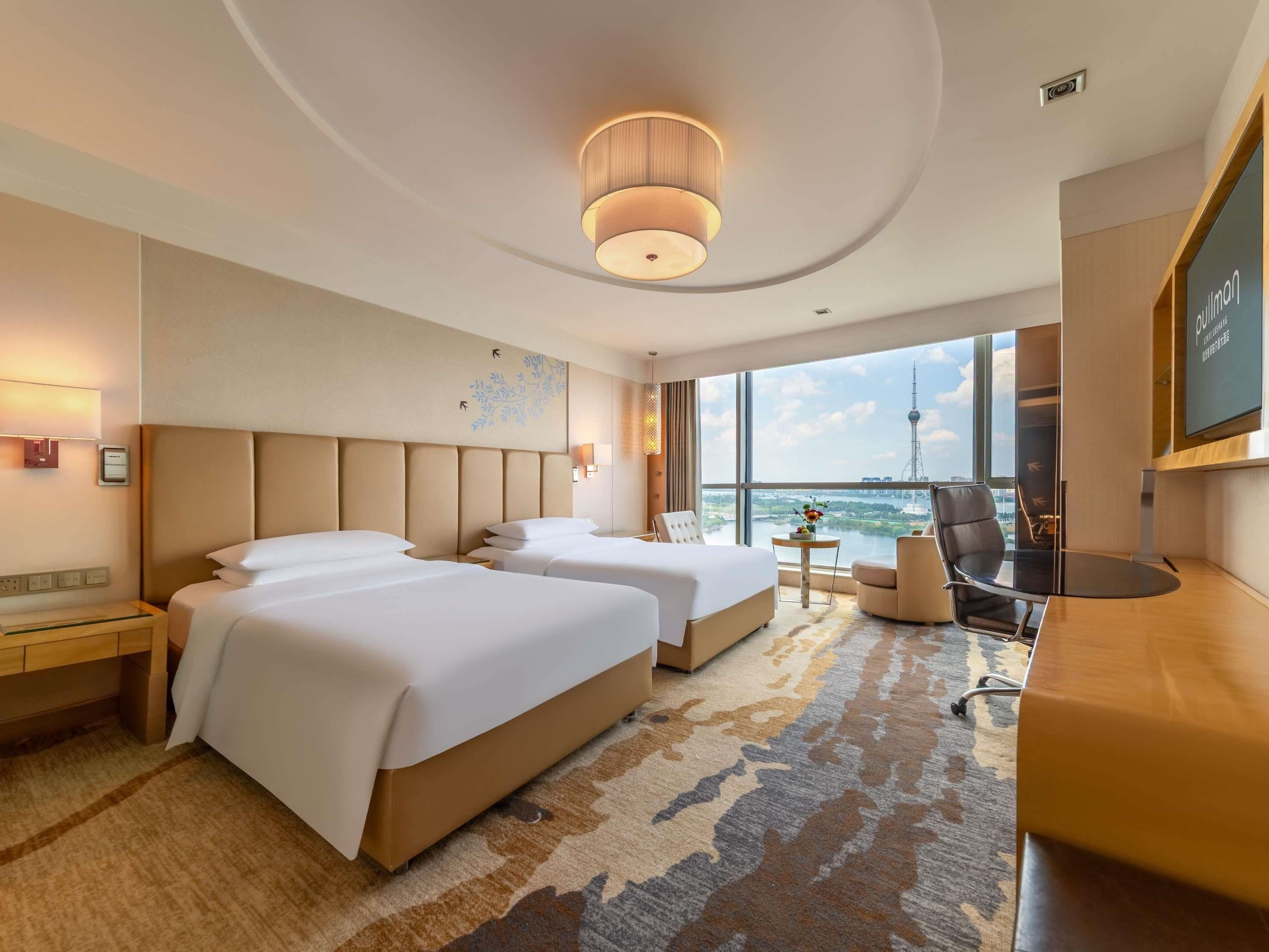 Habitación de hotel con dos camas, un escritorio, una silla y vistas a un paisaje urbano, además de un ventanal grande.