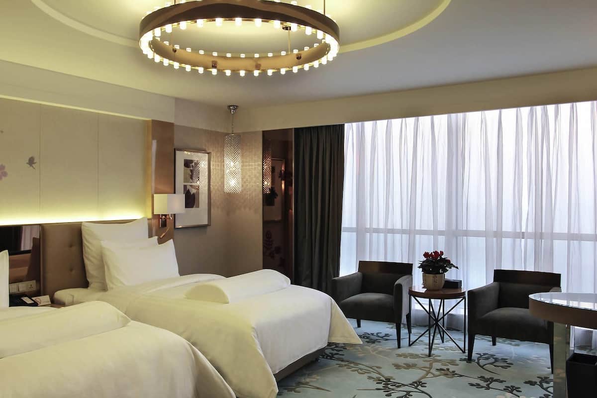 Ett hotellrum med två sängar, en kristallkrona, ett litet bord med en vas och ett stort fönster med tunna, genomskinliga gardiner.