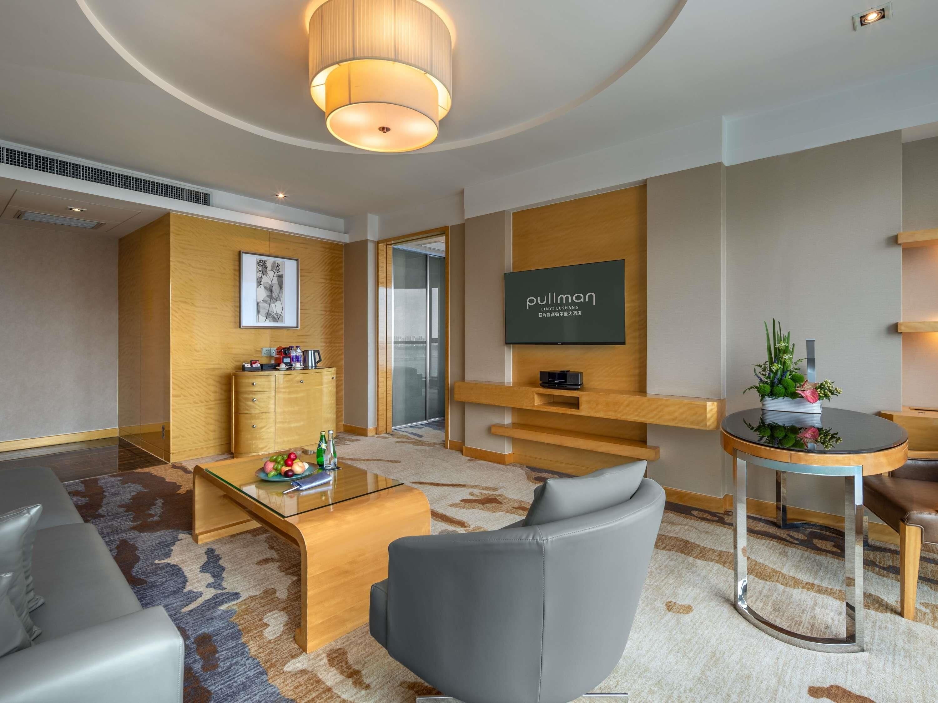 Una habitación de hotel moderna con televisión de pantalla plana, un mueble de madera, una mesita de centro de vidrio, un sofá gris y una mesita auxiliar redonda con un arreglo floral.