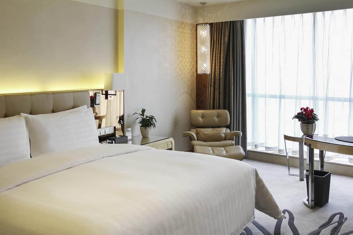 Ett modernt hotellrum med en stor säng, en sittgrupp med en fåtölj och ett bord samt ett stort fönster med gardiner.