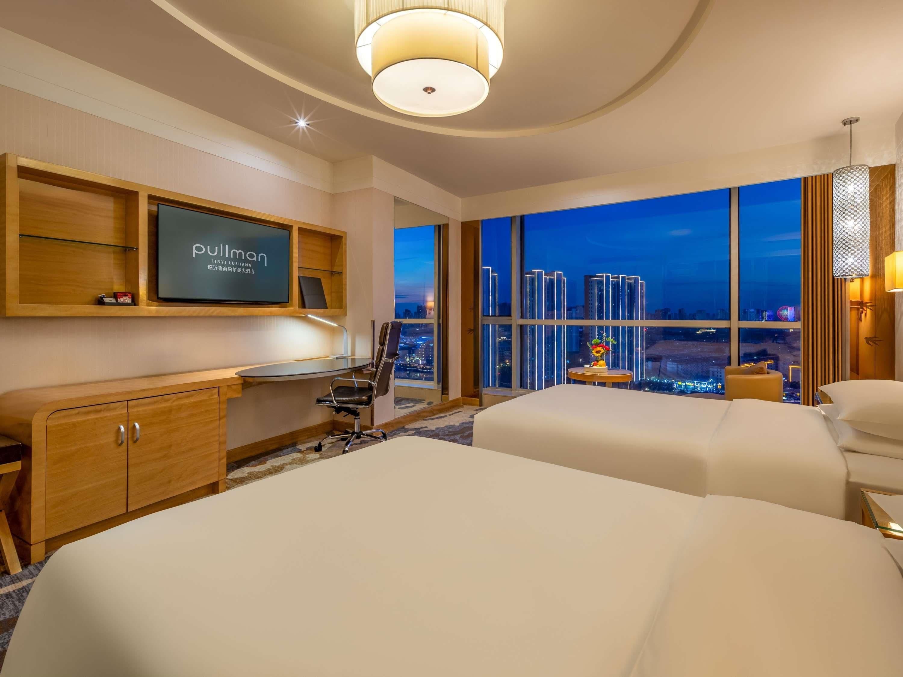 A hotel room with a desk, two beds, a TV, and a city view.