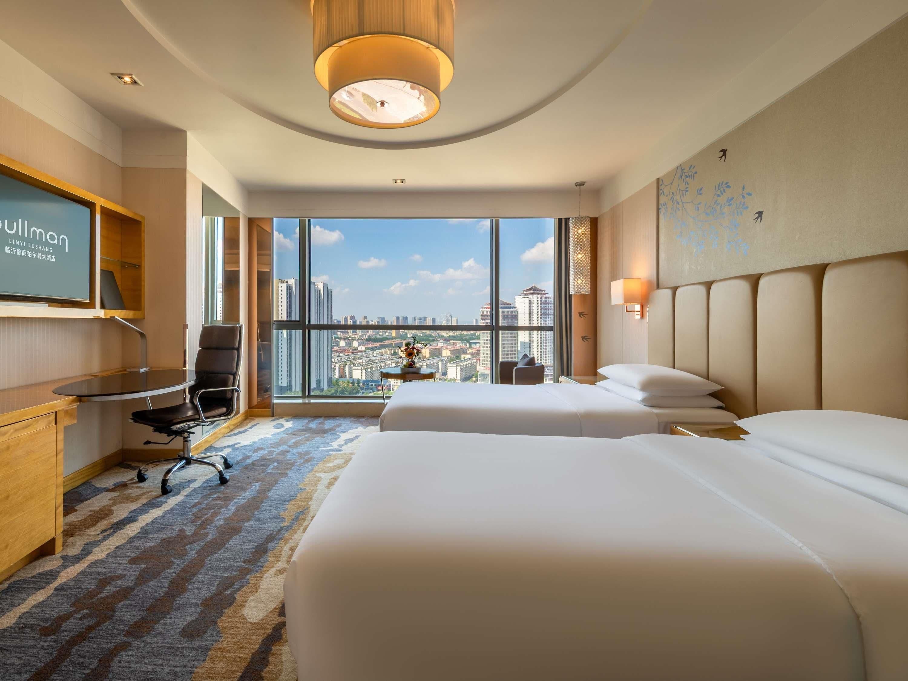 A hotel room with two beds, a desk, a chair, and a large window offering a city view.
