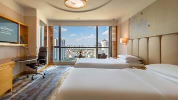 A hotel room with two beds, a desk, a chair, and a large window offering a city view.
